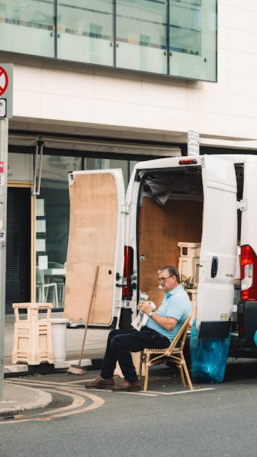 A man wearing glasses and a light blue shirt is seated on a wooden chair, resting outside a white commercial van parked on the street in front of a modern building with large glass windows. The open rear doors of the van reveal a spacious interior filled with packed cardboard boxes and wrapped furniture items, indicating a home relocation process. Several large cardboard panels and a small wooden pallet are set beside the van, suggesting ongoing loading or unloading activities. The man is holding a smartphone or small device, appearing to be taking a break or coordinating the move. The surrounding environment includes a pavement with road markings and a traffic sign partially visible at the top left corner, reflecting an urban setting typical of professional removals services provided by Man with Van Brent, specializing in furniture transport, packing, and moving logistics near Brent and Kilburn High Road.