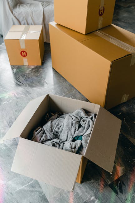 A cardboard box filled with crumpled grey packing paper and clothing, placed on a dark, textured floor inside a home, with several other sealed cardboard boxes of various sizes around it, some marked with shipping labels and tapes. The scene captures the packing stage of a home relocation process, with boxes ready for loading onto a van for a professional house removal service. The lighting is natural, highlighting the different materials used for packing, such as cardboard, fabric, and paper, emphasizing the careful and organized nature of packing and moving. This setup reflects the typical preparations involved in furniture transport, packing, and loading prior to a house removal, as managed by companies like Man with Van Brent, operating in the Kilburn High Road area.