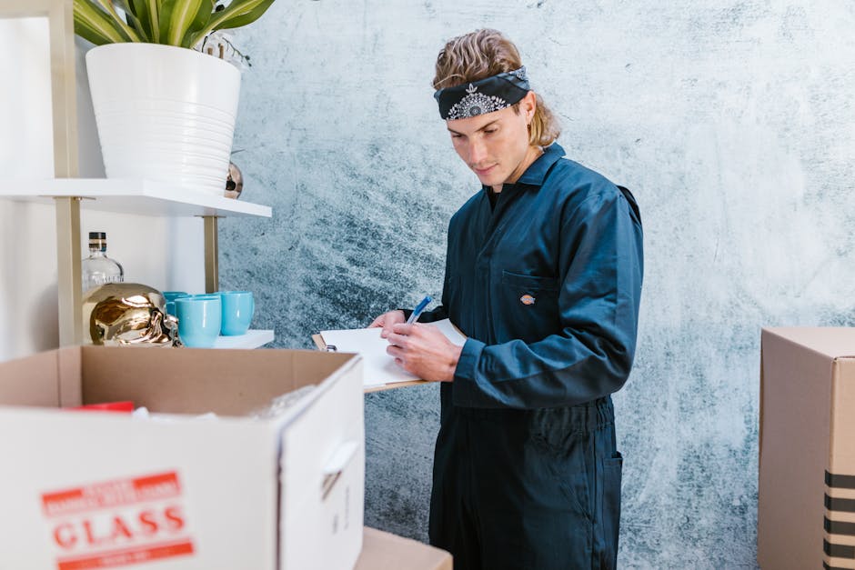 A young man wearing a navy work jumpsuit and a black bandana with white patterns is inside a room with a pale, textured wall. He is holding a clipboard and a pen, appearing to take notes or conduct an