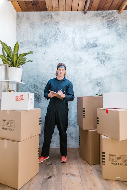 A young man wearing a navy work jumpsuit and a black bandana with white patterns is inside a room with a pale, textured wall. He is holding a clipboard and a pen, appearing to take notes or conduct an